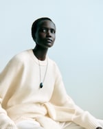 Placeholder for: A person wearing a cream-colored sweater and a pendant necklace, sitting against a plain, light blue background.