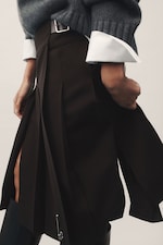 Placeholder for: Person wearing a dark pleated skirt with a belt, a gray sweater with white cuffs, and black boots, holding the skirt with one hand.