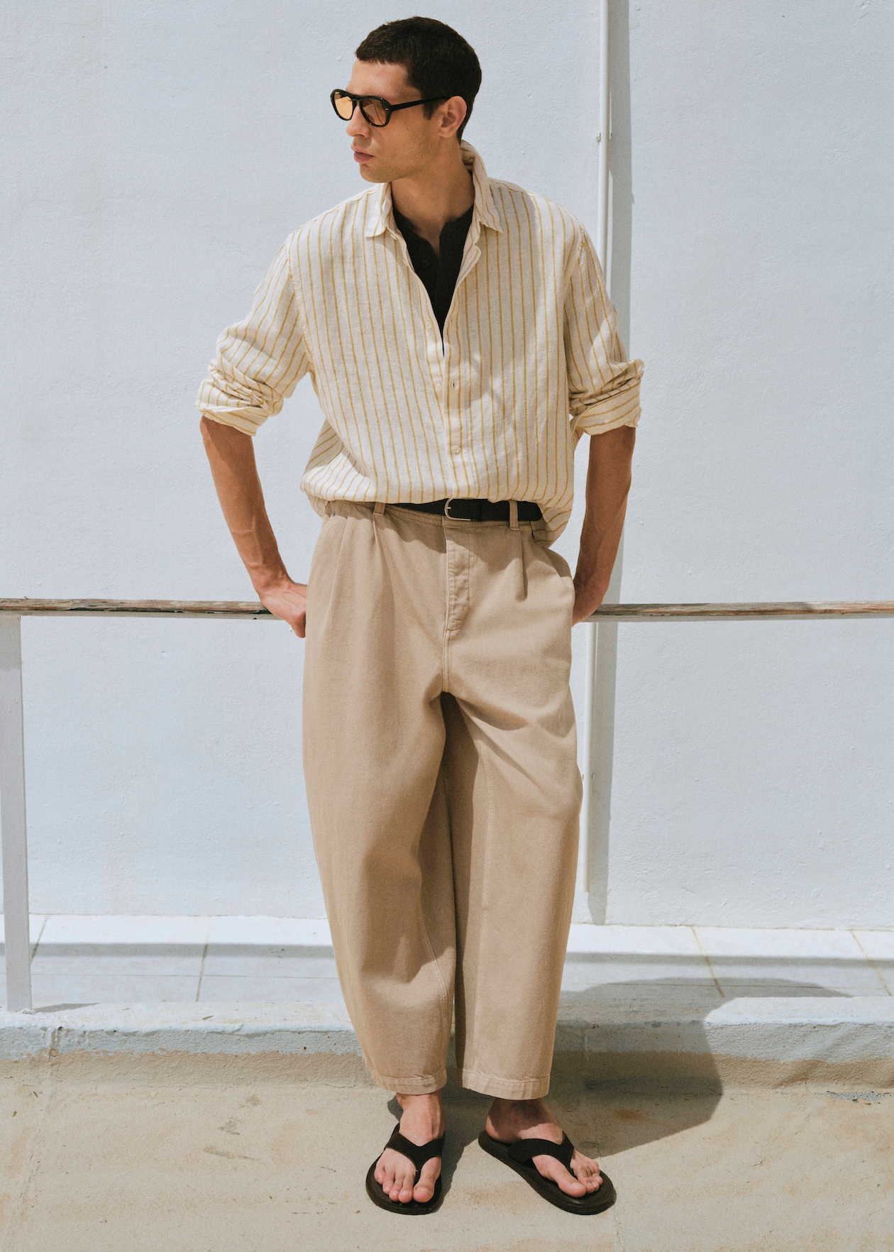Man in a striped shirt, beige trousers, and flip-flops leans against a railing, looking to the side, with a light blue background.
