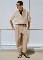 Placeholder for: Man in a striped shirt, beige trousers, and flip-flops leans against a railing, looking to the side, with a light blue background.
