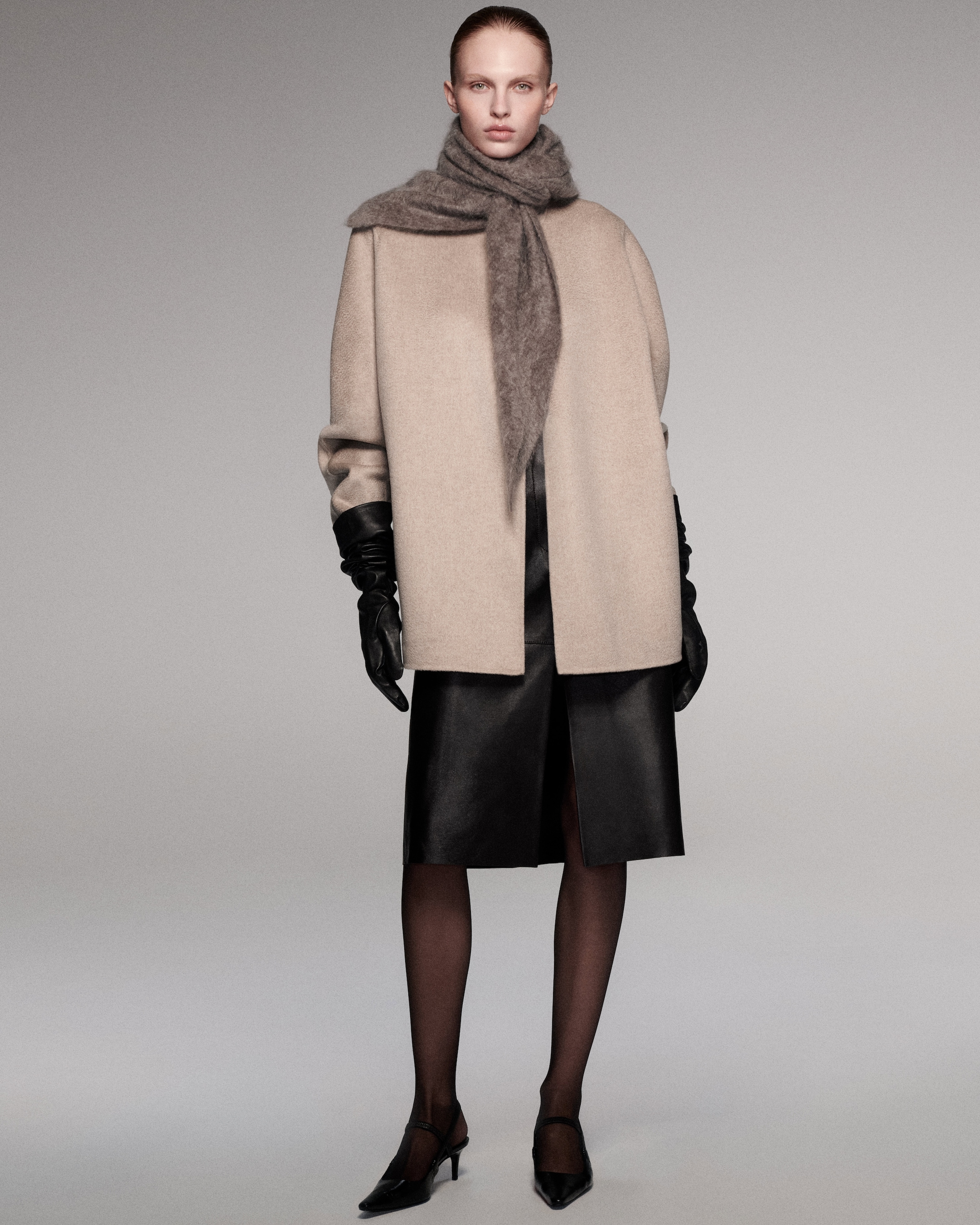 A model wearing a beige coat and scarf, with black gloves, skirt, tights, and heels, standing against a gray background.