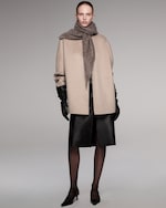 Placeholder for: A model wearing a beige coat and scarf, with black gloves, skirt, tights, and heels, standing against a gray background.
