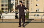Placeholder for: Man in sunglasses wearing a dark matching outfit stands on a rooftop, with arms outstretched, cityscape in the background.
