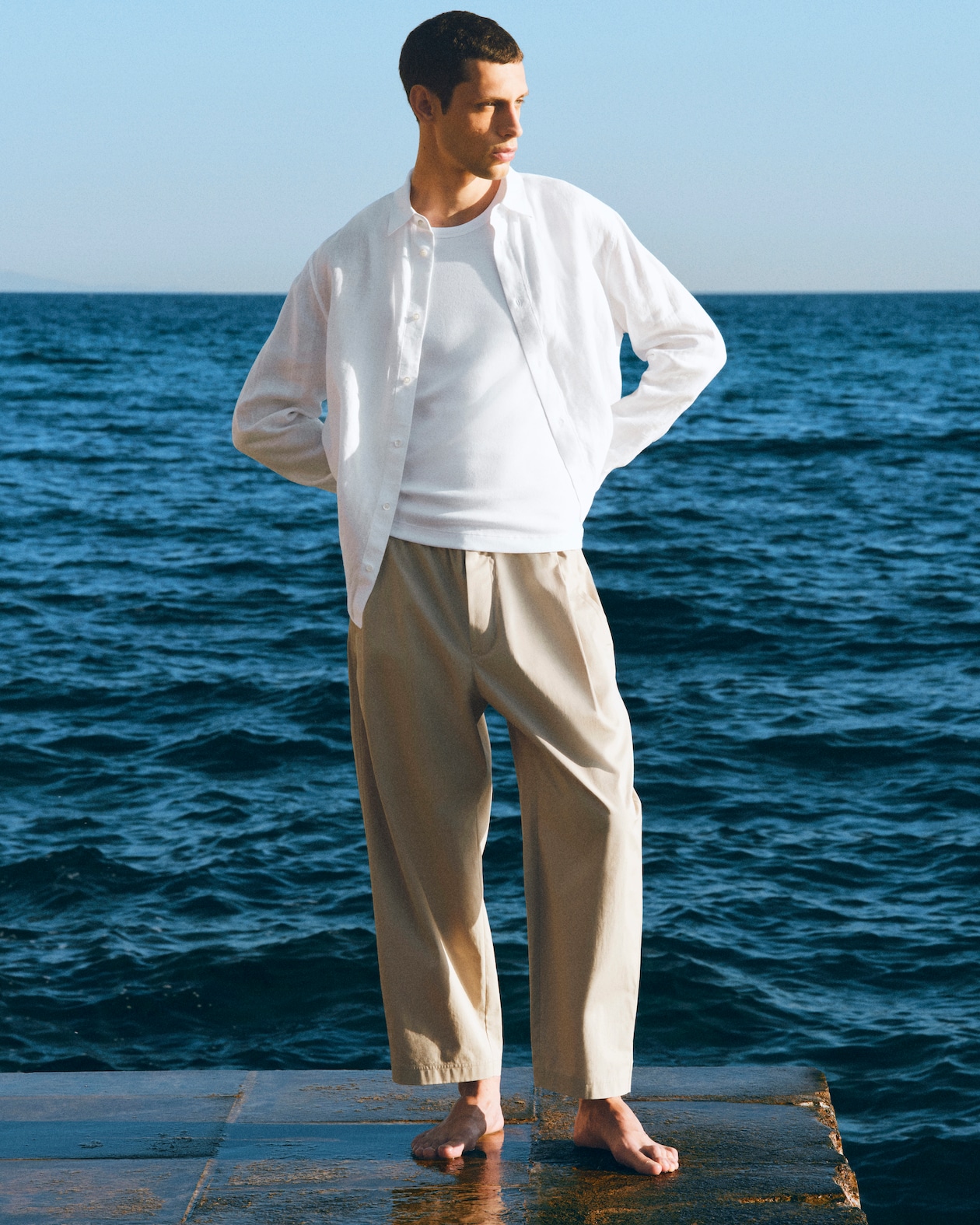 A person in a white shirt and beige pants stands barefoot on a wet surface by the sea, looking into the distance.