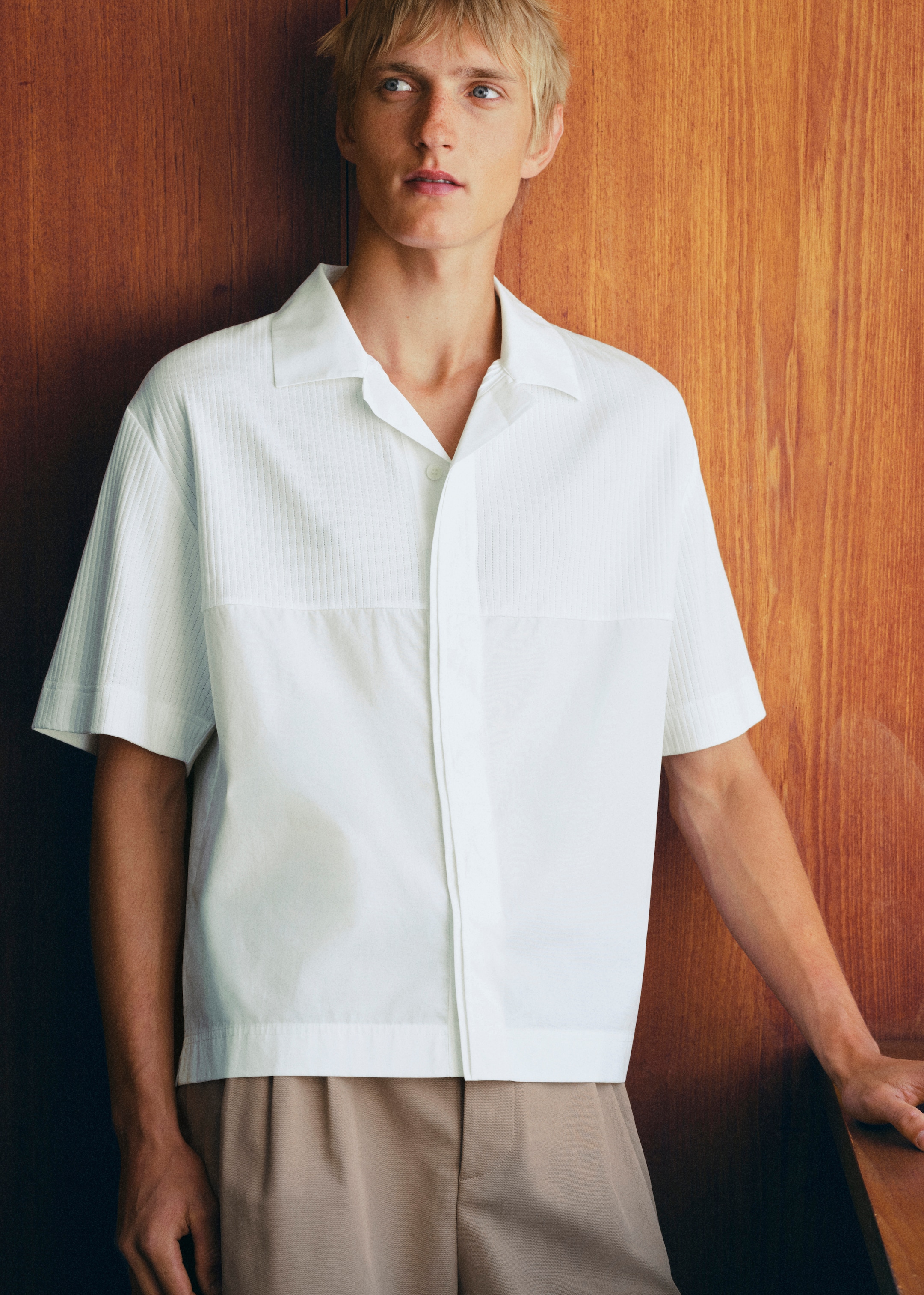 A person with short blond hair wearing a white shirt and beige pants stands against a wooden wall, looking off to the side.