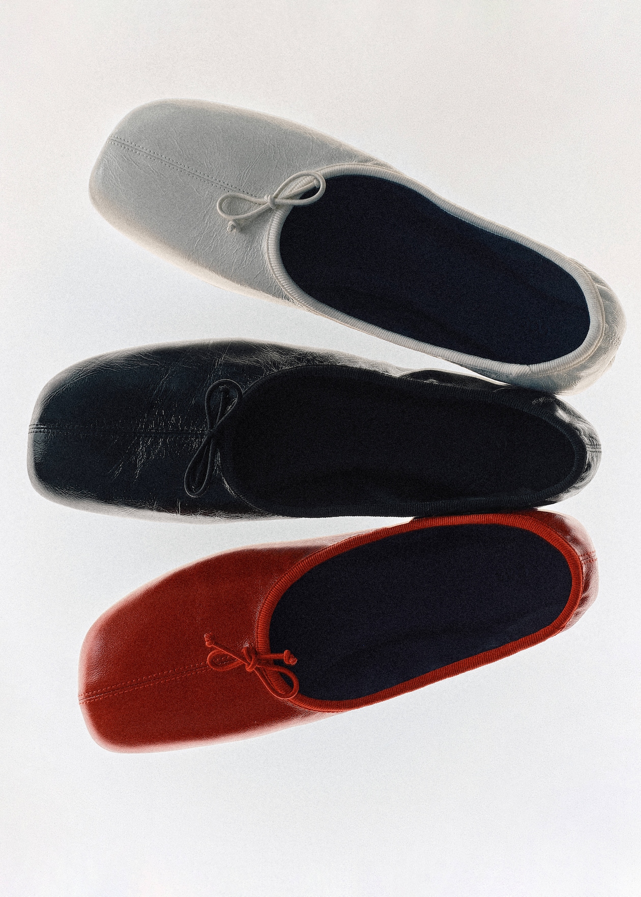 Three pairs of ballet flats in white, black, and red, arranged vertically with bows on the toes against a plain background.