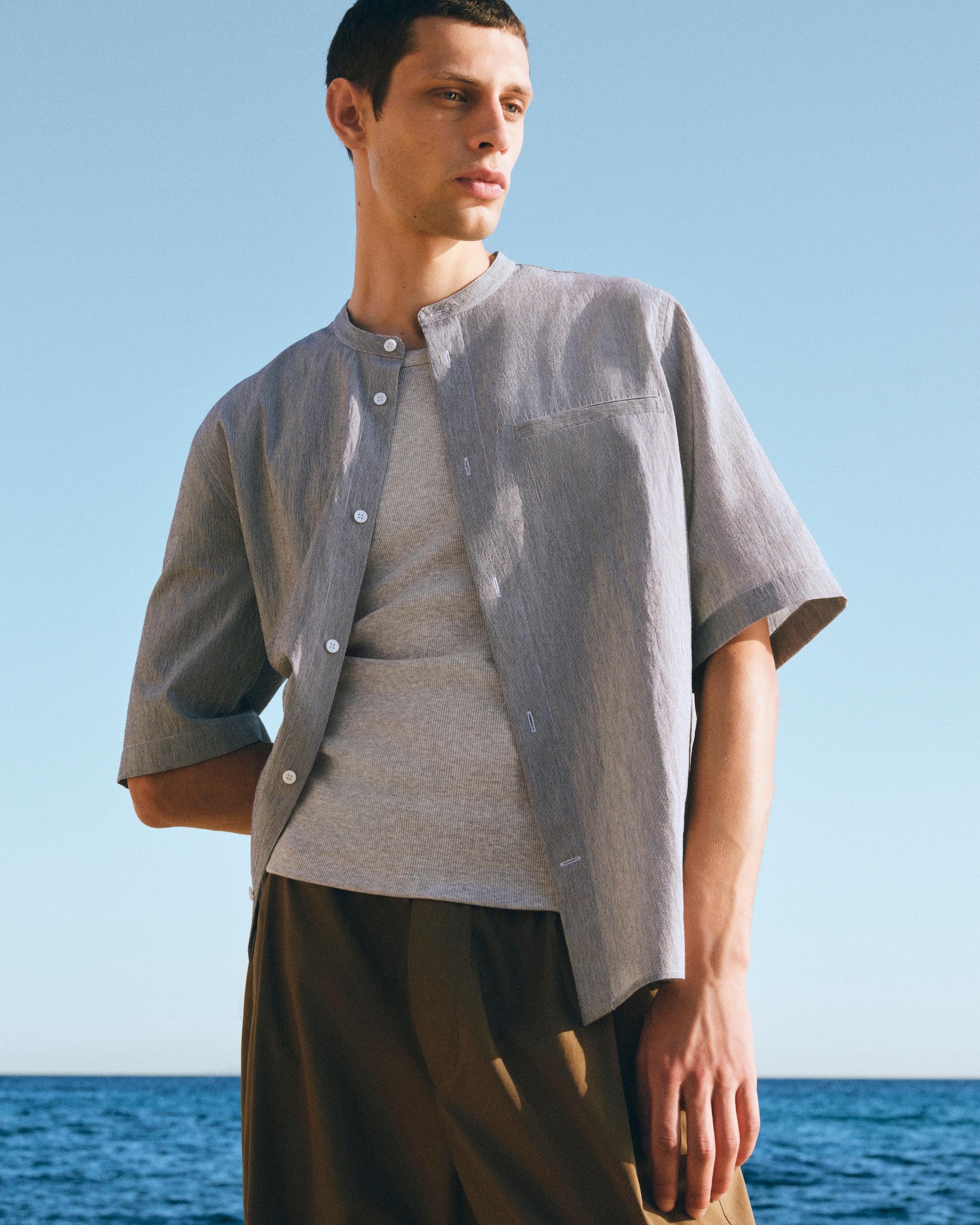 Man in a grey button-up shirt and brown pants standing by the sea, looking into the distance on a clear day.