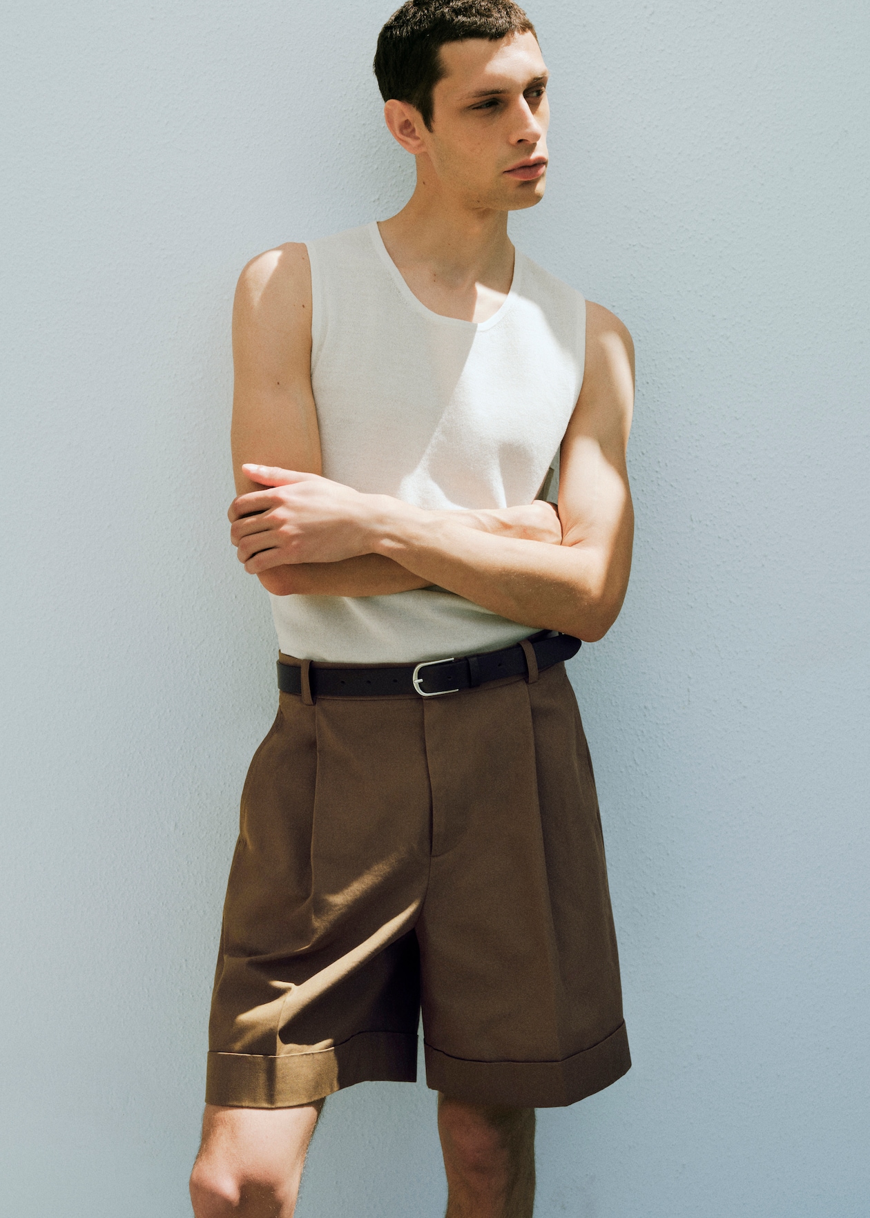 Man in a white sleeveless top and brown shorts, standing against a plain white wall with arms crossed.