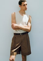 Placeholder for: Man in a white sleeveless top and brown shorts, standing against a plain white wall with arms crossed.