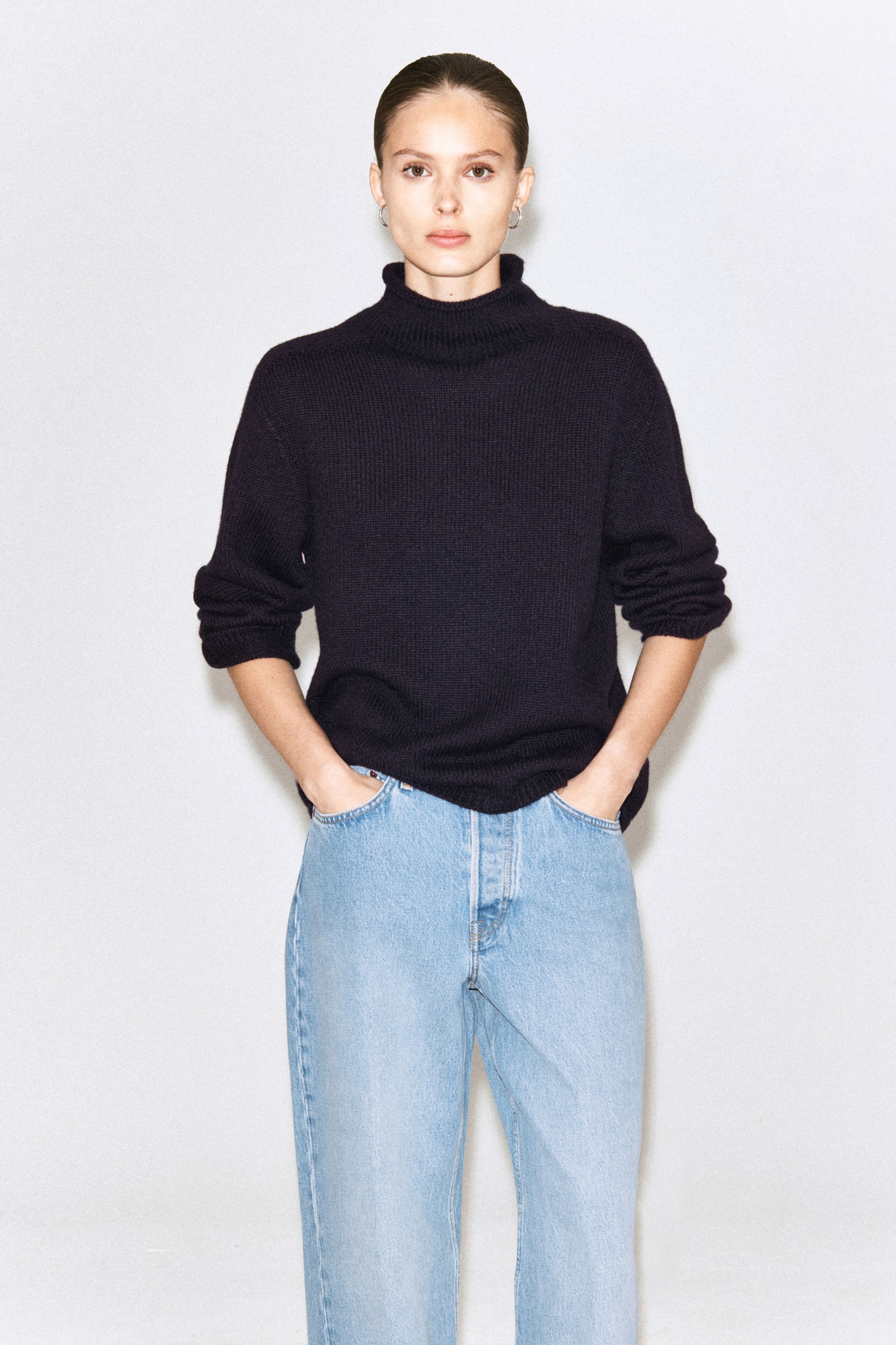 A person in a black turtleneck sweater and light blue jeans stands with hands in pockets against a plain white background.