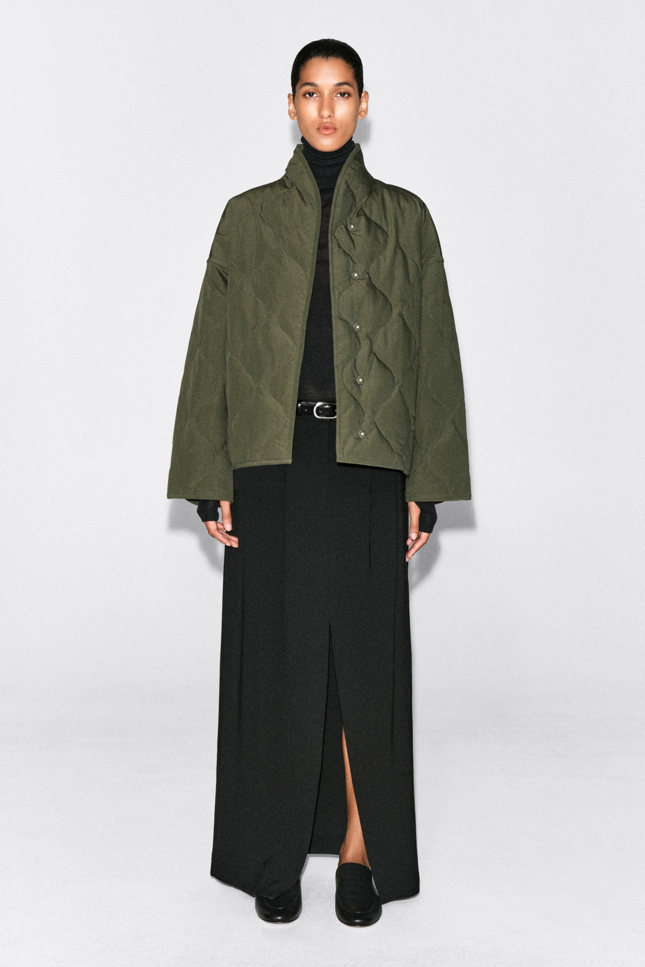 Person in an olive green quilted jacket over a black turtleneck and long black skirt, standing against a plain white background.