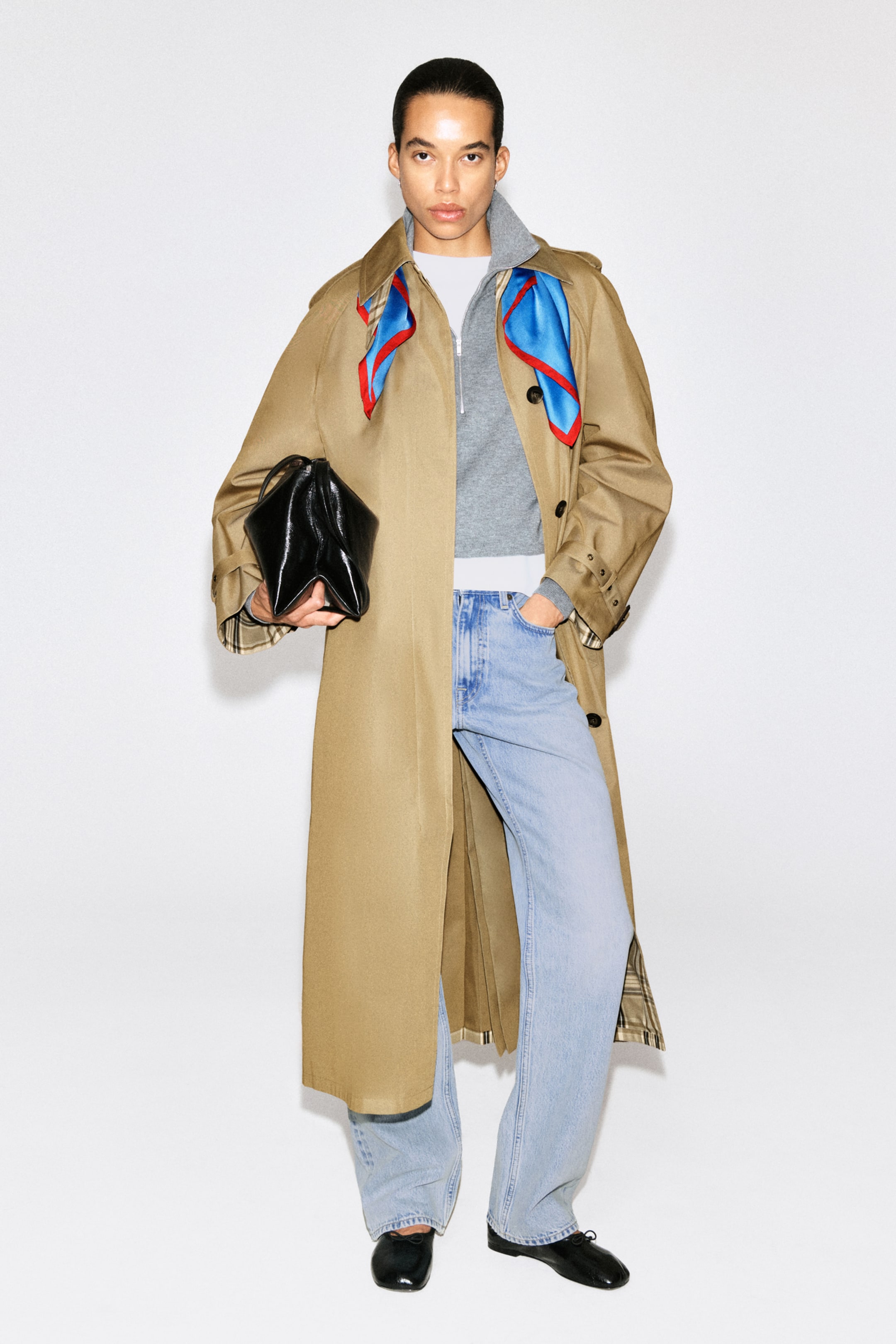 A person wearing a long beige trench coat, blue jeans, and black shoes, holding a black handbag. A colorful scarf is draped around the neck.