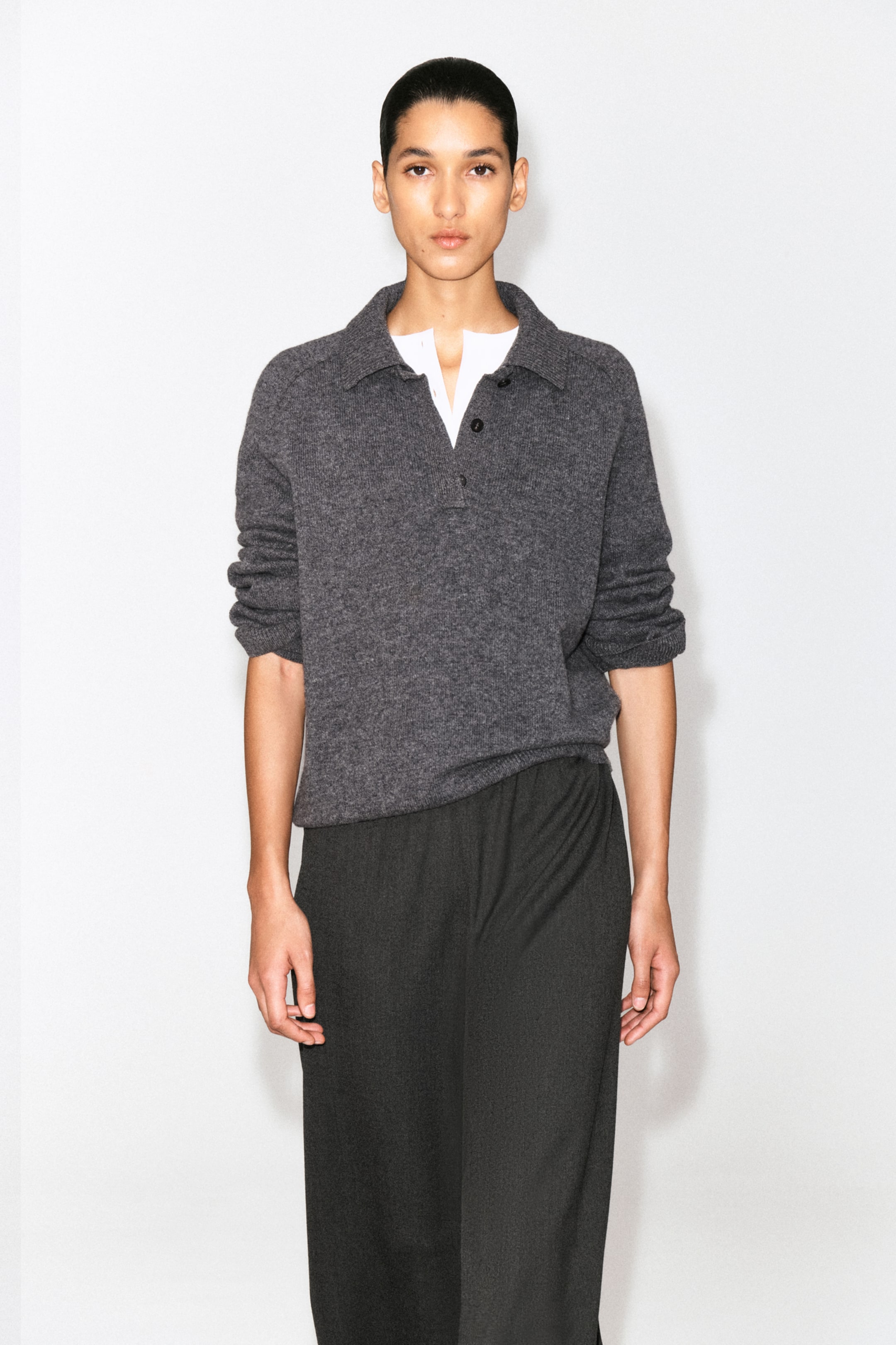 Person wearing a grey sweater over a white shirt and dark trousers, standing against a plain white background.