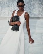 Placeholder for: Person in a white sleeveless dress, holding a black bag, wearing sunglasses, and leaning against a textured wall.