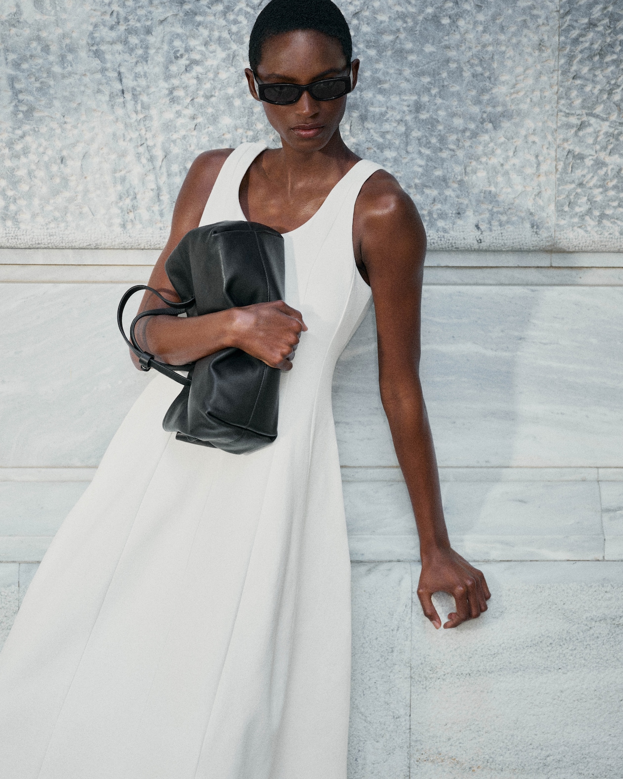A person in a white sleeveless dress, wearing sunglasses, leans against a wall, holding a black handbag.