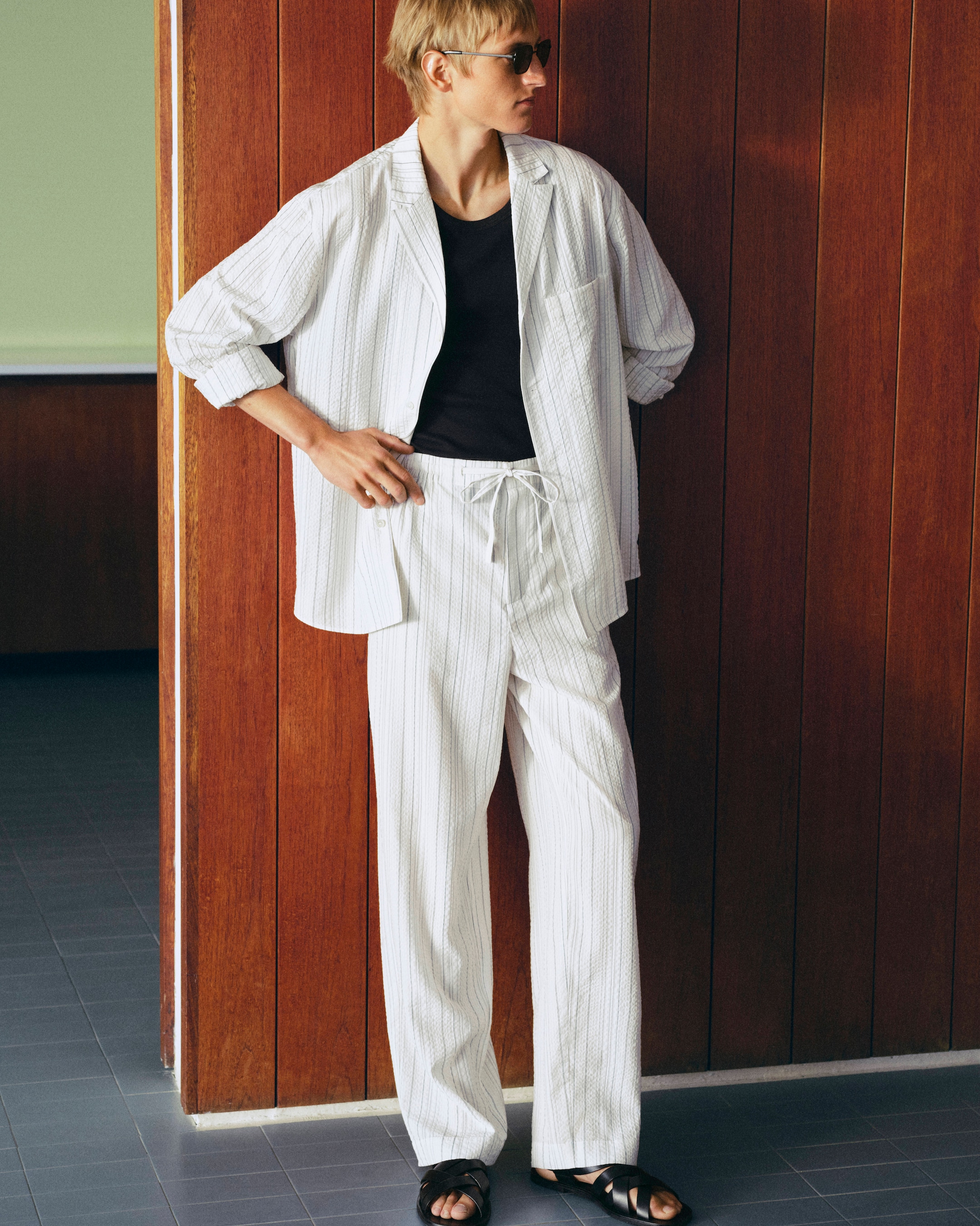 Person in sunglasses wearing a casual white pinstripe outfit over a black shirt, leaning against a wooden wall.