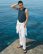 Placeholder for: Man in a navy tank top and white pants stands on a seaside stone dock, gazing at the ocean. A white shirt is tied around his waist.