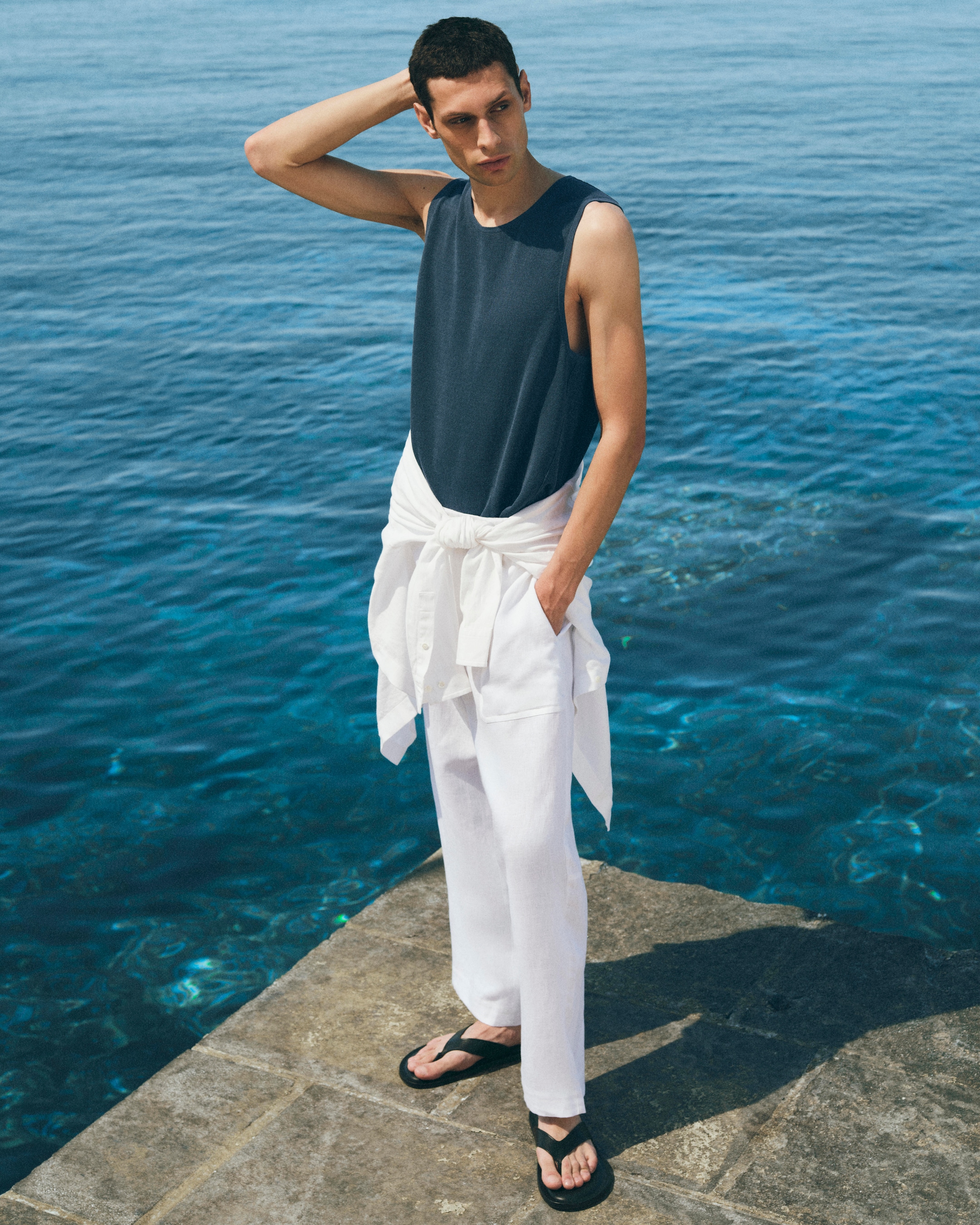 Man in a dark tank top and white pants stands on a pier by the sea, looking relaxed with one hand on his head.