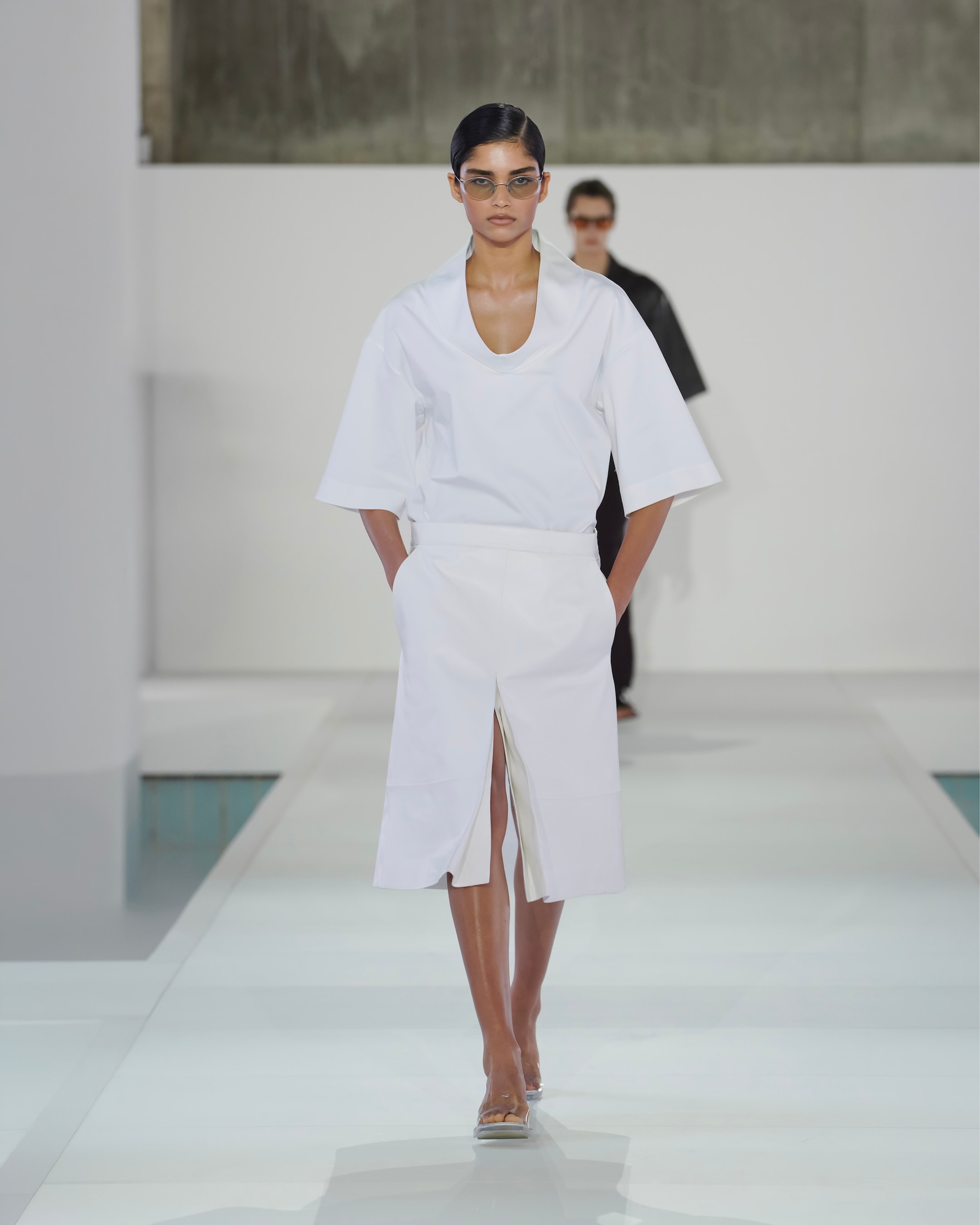 Model in a white outfit with a V-neck top and skirt walks confidently on a runway; wearing sunglasses, sleek hairstyle.