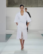 Placeholder for: Model in a white outfit with a V-neck top and skirt walks confidently on a runway; wearing sunglasses, sleek hairstyle.