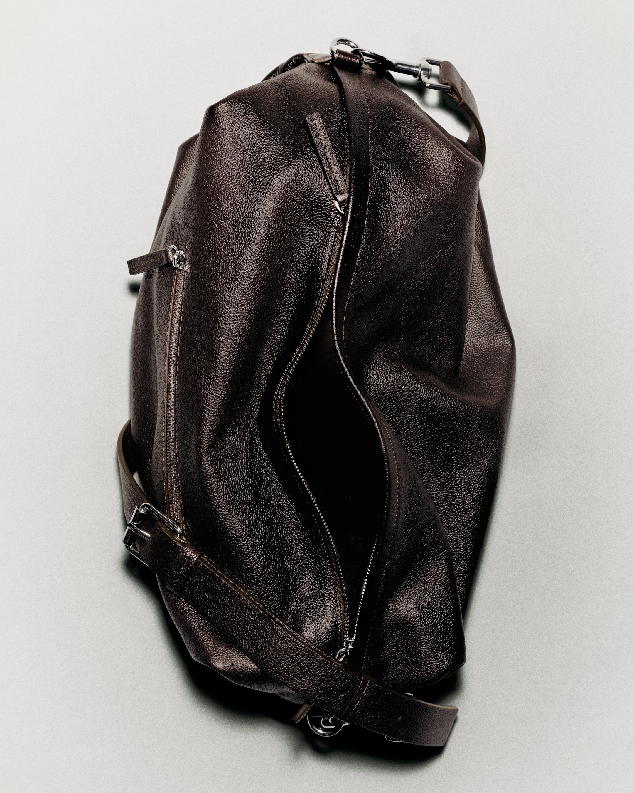 A brown leather duffel bag with zippers and adjustable strap, slightly open, lying on a light gray surface.