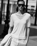Placeholder for: Black and white photo of a person wearing sunglasses and a light-colored, short-sleeve top, holding a jacket, walking outside.