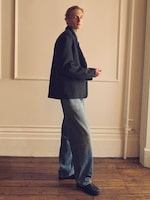Placeholder for: Model in an oversized dark blazer and wide-legged jeans standing on a wooden floor, with a neutral wall in the background.