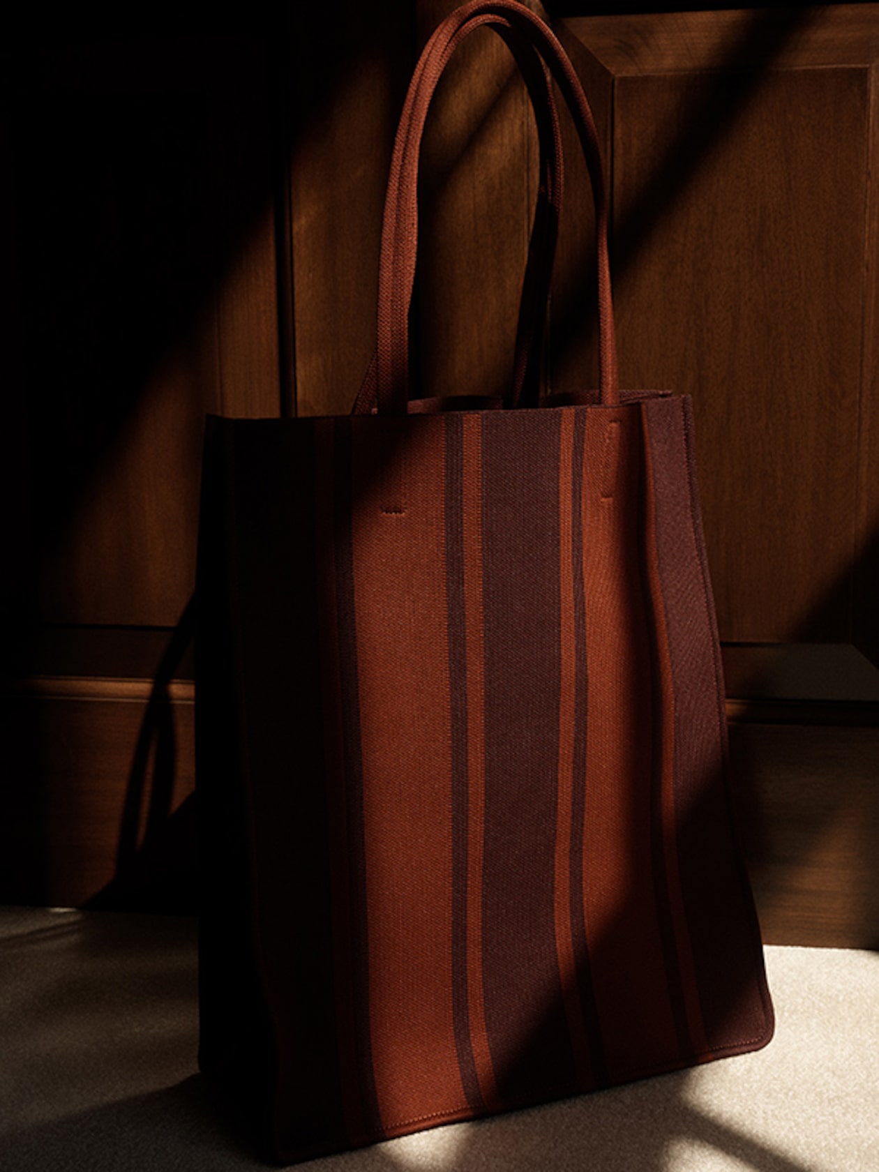 A stylish tote bag with vertical maroon and dark red stripes, placed against a wooden background with dramatic lighting and shadows.