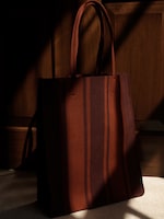 Placeholder for: A stylish tote bag with vertical maroon and dark red stripes, placed against a wooden background with dramatic lighting and shadows.