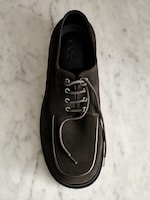 Placeholder for: Top view of a single brown suede shoe with grey laces on a white marble surface. The brand name is visible inside the shoe.