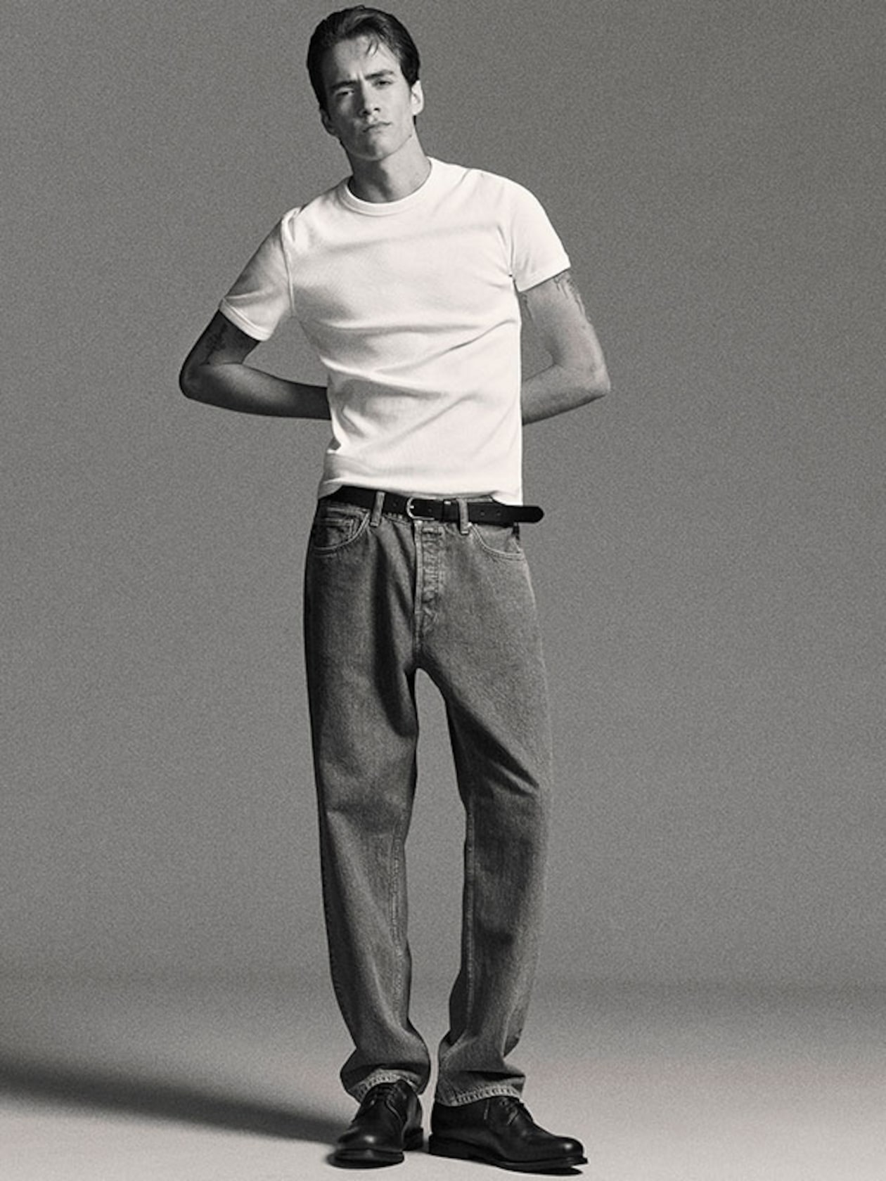 Model wearing a white T-shirt, relaxed jeans, and black shoes stands with hands behind his back against a plain background.