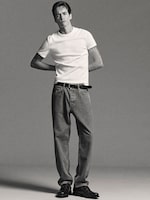 Placeholder for: Model wearing a white T-shirt, relaxed jeans, and black shoes stands with hands behind his back against a plain background.