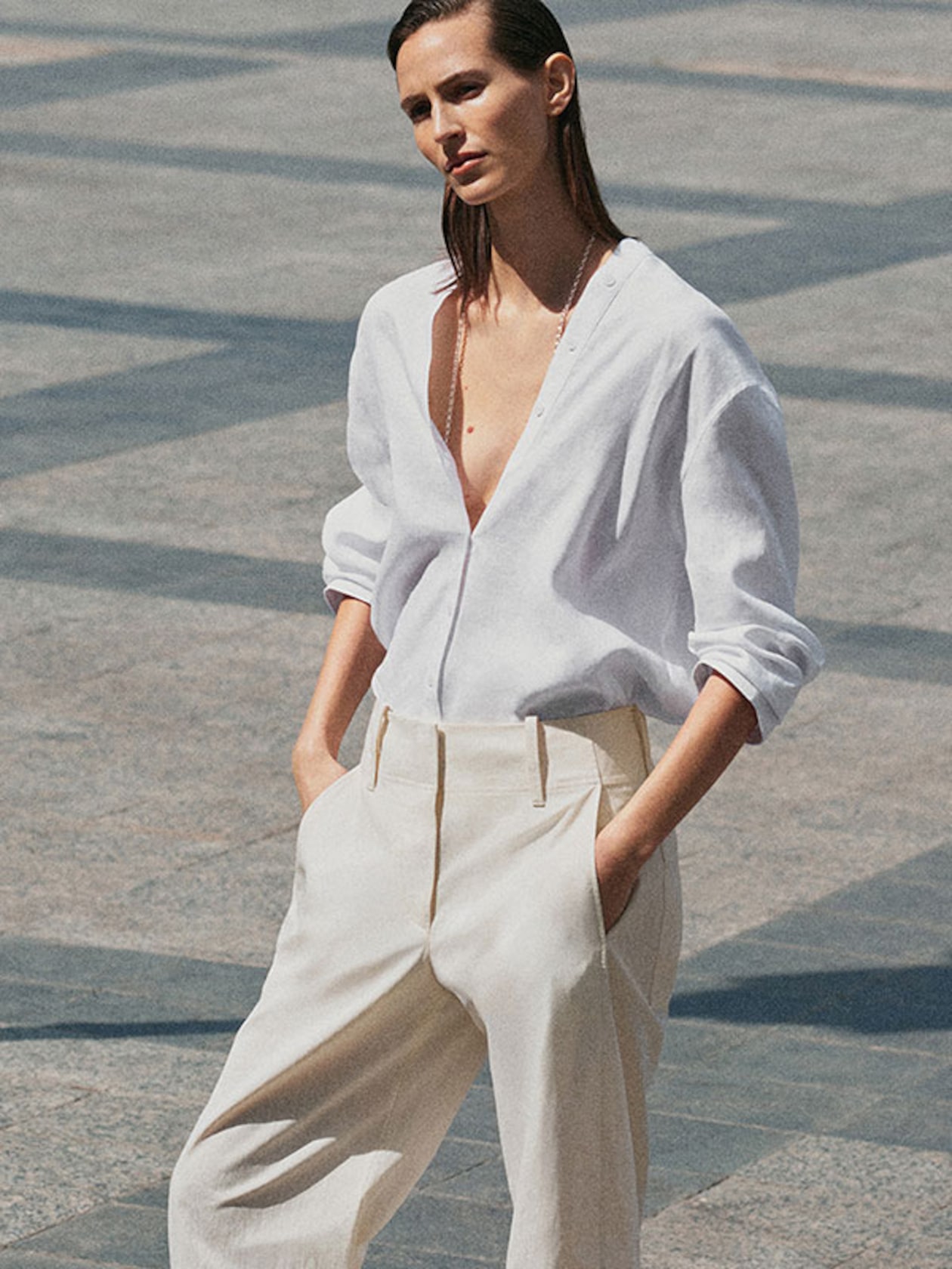 A person in a white shirt and beige pants stands confidently, hands in pockets, against a tiled outdoor backdrop.