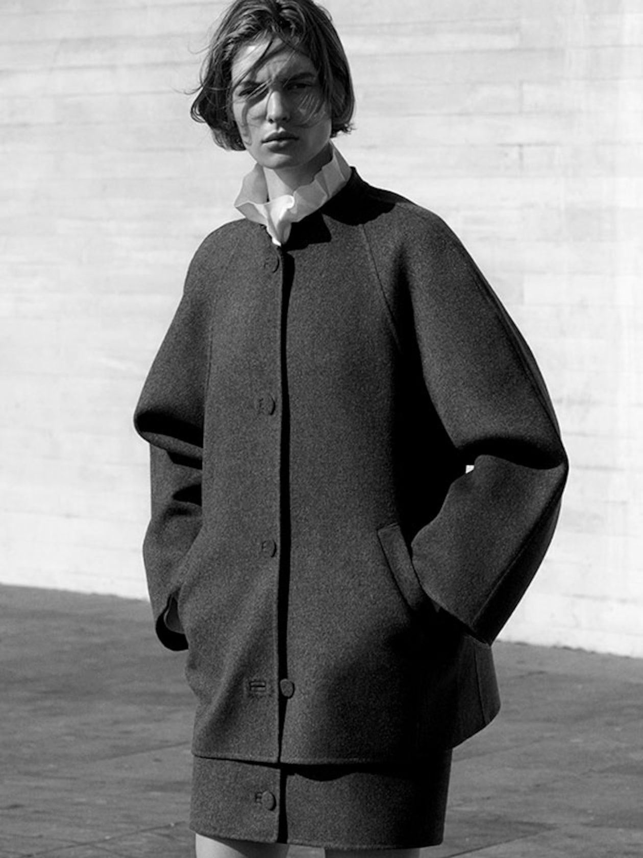 Black and white photo of a person in a large coat and white blouse standing against a light-colored wall, looking at the camera.
