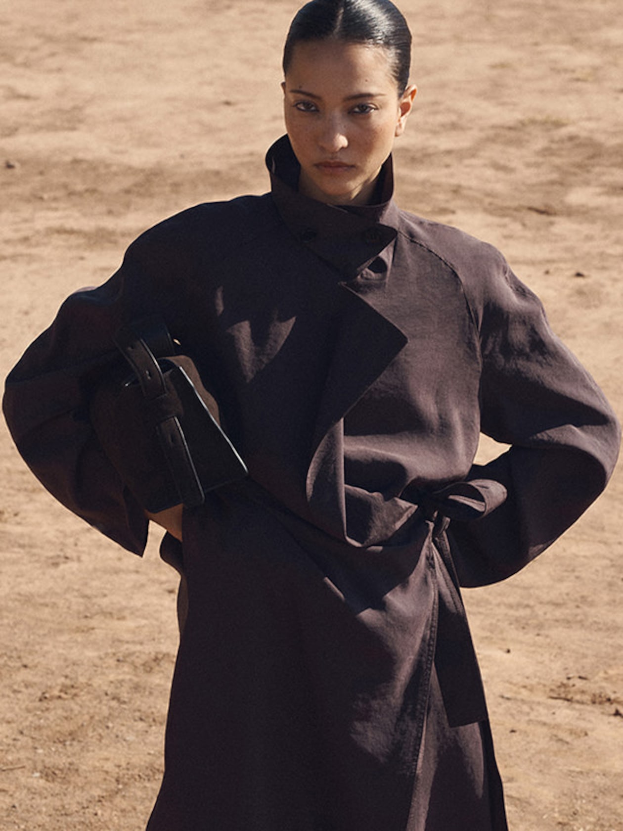 A person in a dark trench coat stands confidently in a sandy outdoor setting, holding a matching handbag under one arm.