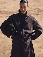 Placeholder for: A person in a dark trench coat stands confidently in a sandy outdoor setting, holding a matching handbag under one arm.