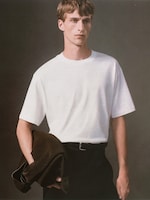 Placeholder for: Person in a white T-shirt and black pants holds a dark jacket against a neutral background.