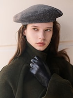 Placeholder for: A person in a dark coat and black gloves wears a gray beret, looking contemplative against a light-colored background.