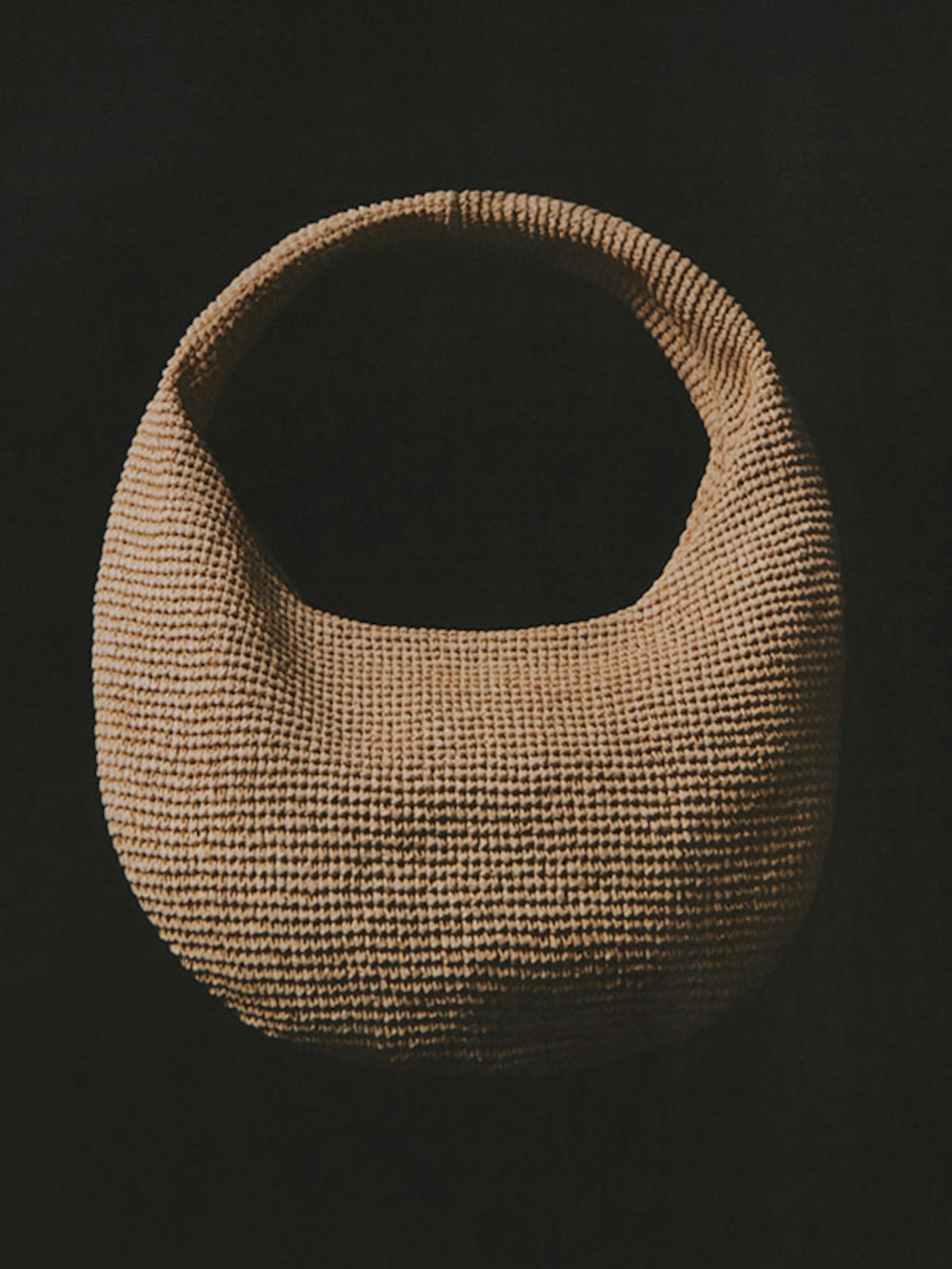 A woven, tan-colored, crescent-shaped bag with a wide handle, set against a black background.