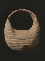 Placeholder for: A woven, tan-colored, crescent-shaped bag with a wide handle, set against a black background.
