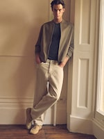 Placeholder for: Person in a casual outfit leans against a wall by a window, wearing a brown jacket, black shirt, beige pants, and sneakers.