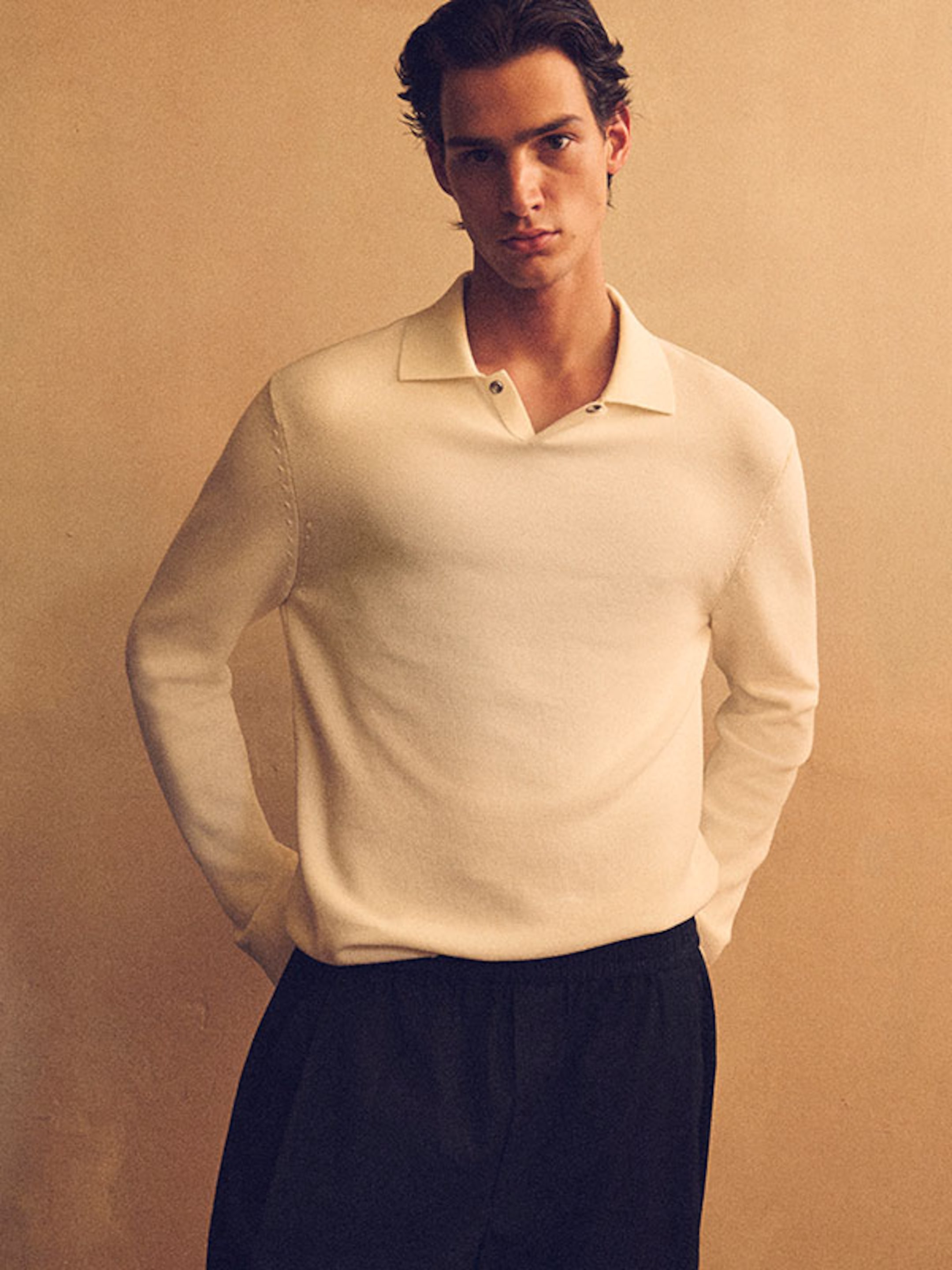 Person wearing a cream-colored sweater and black pants, standing against a beige background with hands in pockets.