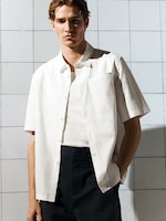 Placeholder for: Model wearing a short-sleeve white shirt over a white tee and dark pants, standing against a tiled wall.