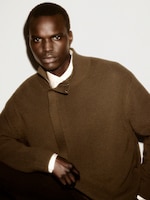 Placeholder for: Person wearing a brown knit sweater and white collared shirt, posing against a plain background, with a serious expression.