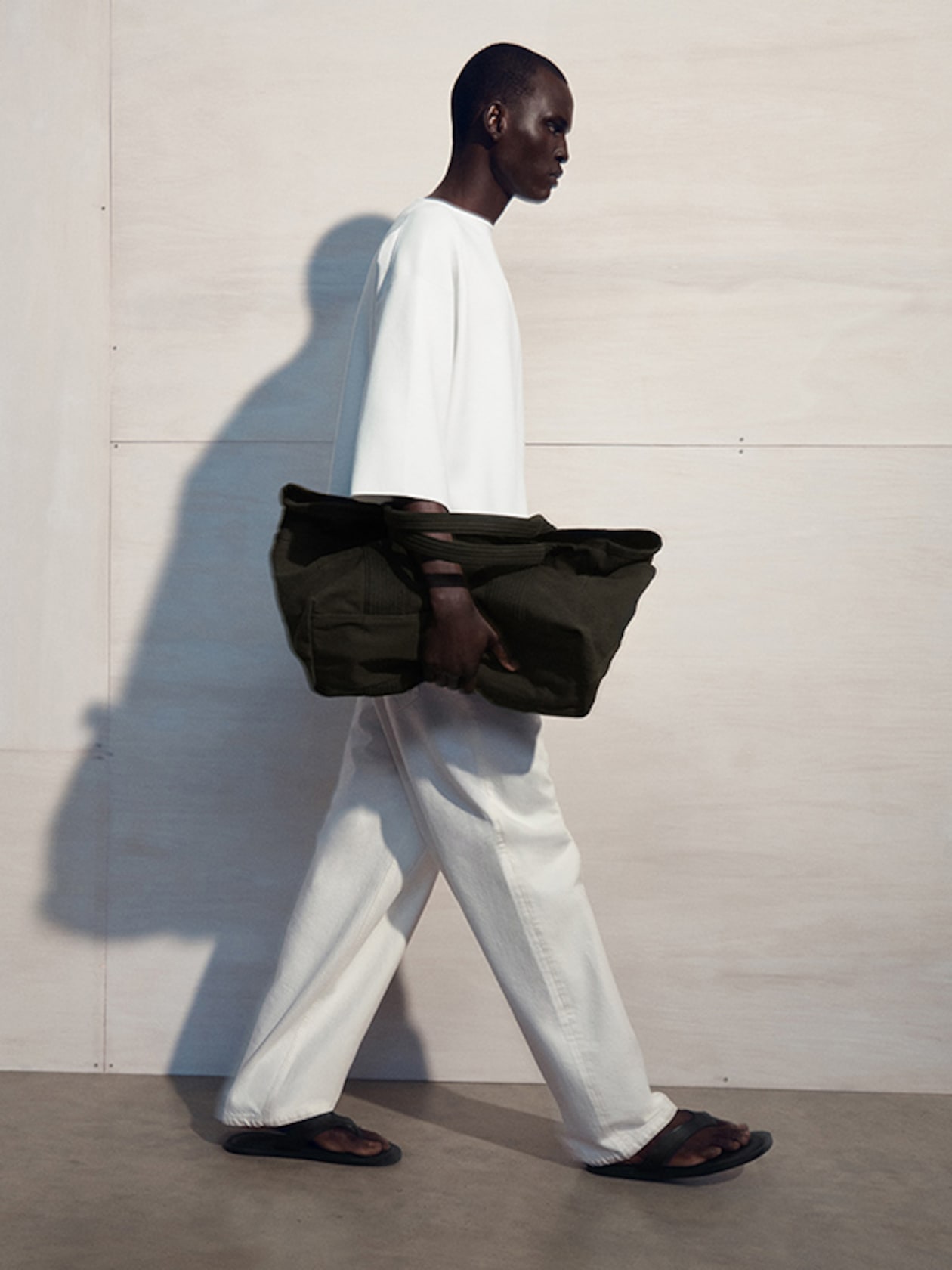 Person in white outfit walking sideways, holding a large dark bag, with a light background and shadow cast on the wall.
