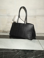 Placeholder for: Black woven handbag with long handles, containing a newspaper, placed on a black-and-white checkered marble floor.