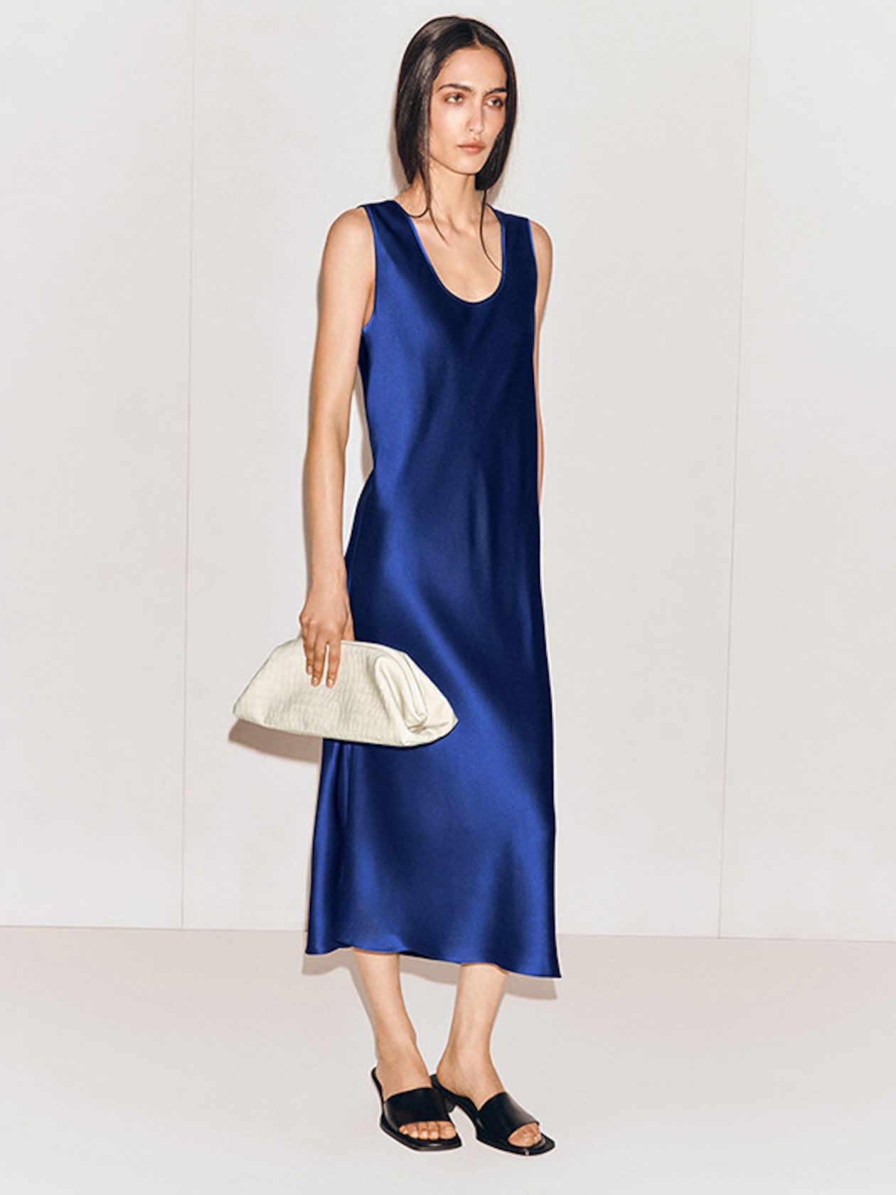 Woman in a sleeveless, blue satin dress holds a white clutch and wears black heeled sandals against a simple background.