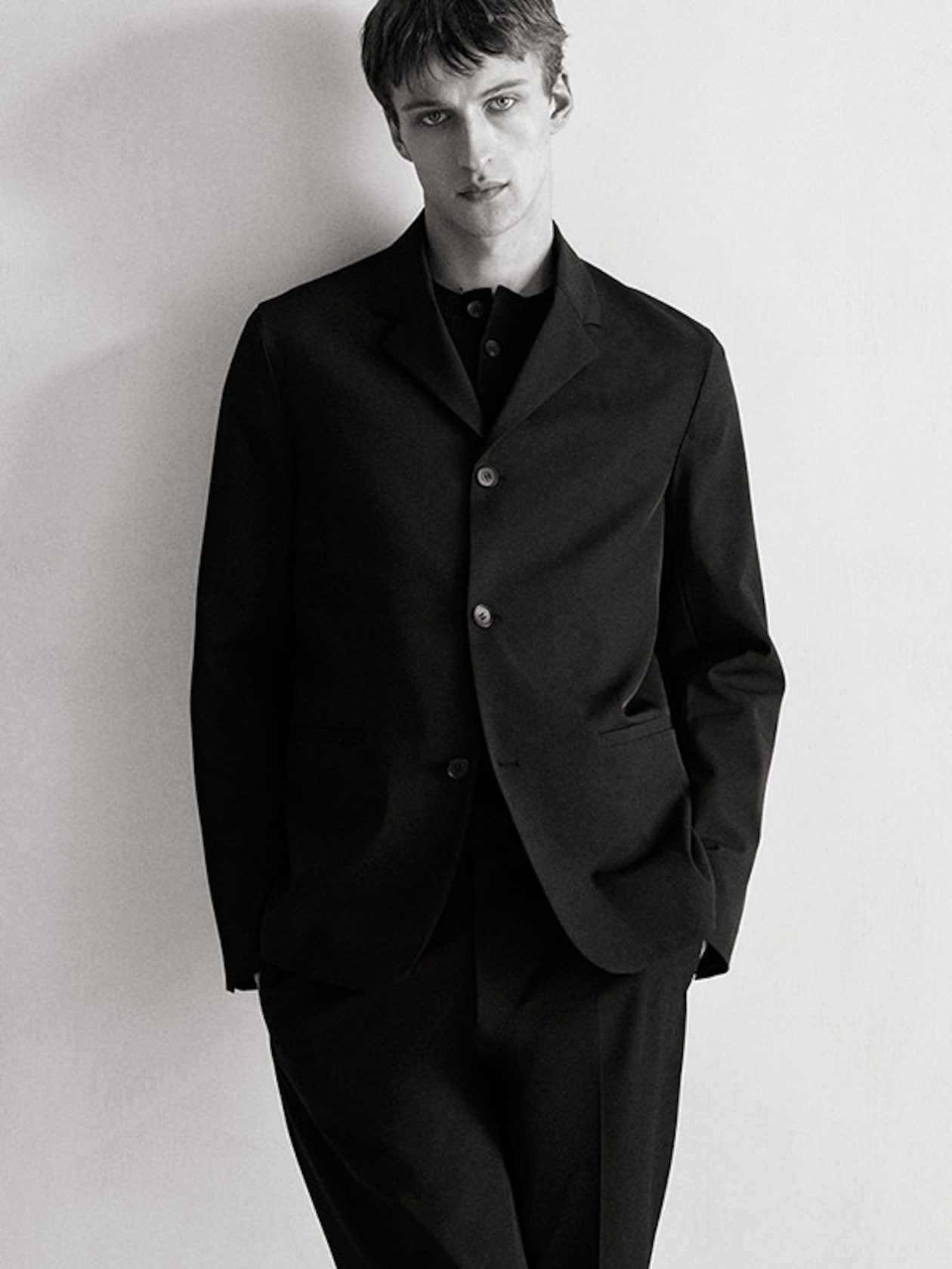 Man in a black suit poses against a plain background, looking directly at the camera with a neutral expression.