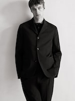 Placeholder for: Man in a black suit poses against a plain background, looking directly at the camera with a neutral expression.