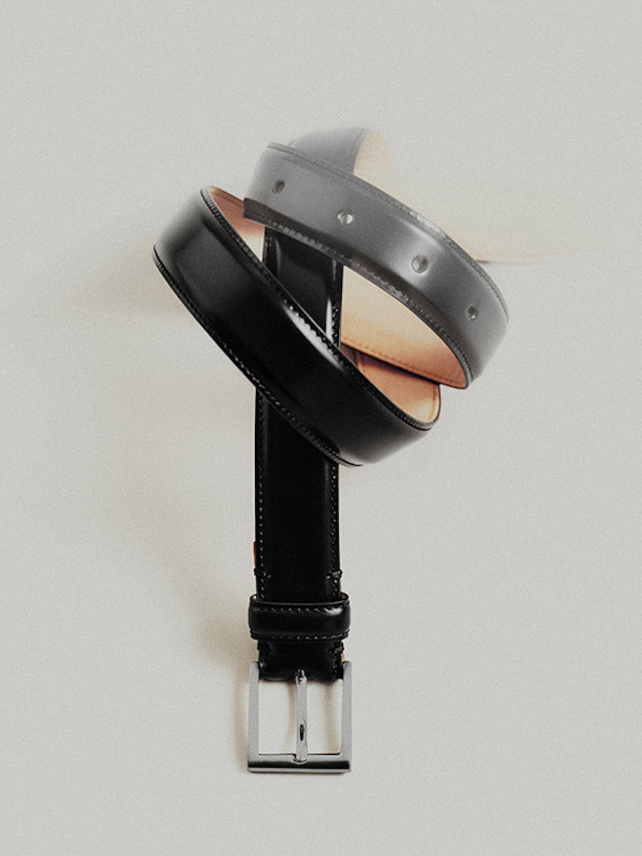 A black leather belt with a silver buckle, artistically twisted into a spiral shape, set against a light gray background.